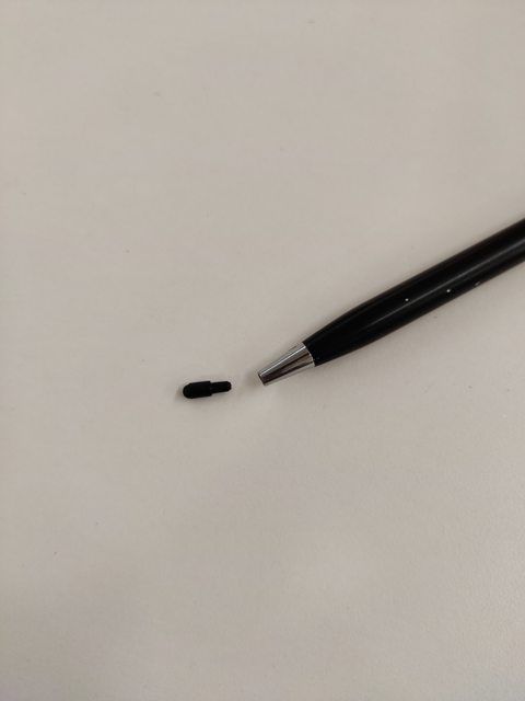 Tiny black micro-injection-moulded part placed next to a pen tip for scale.